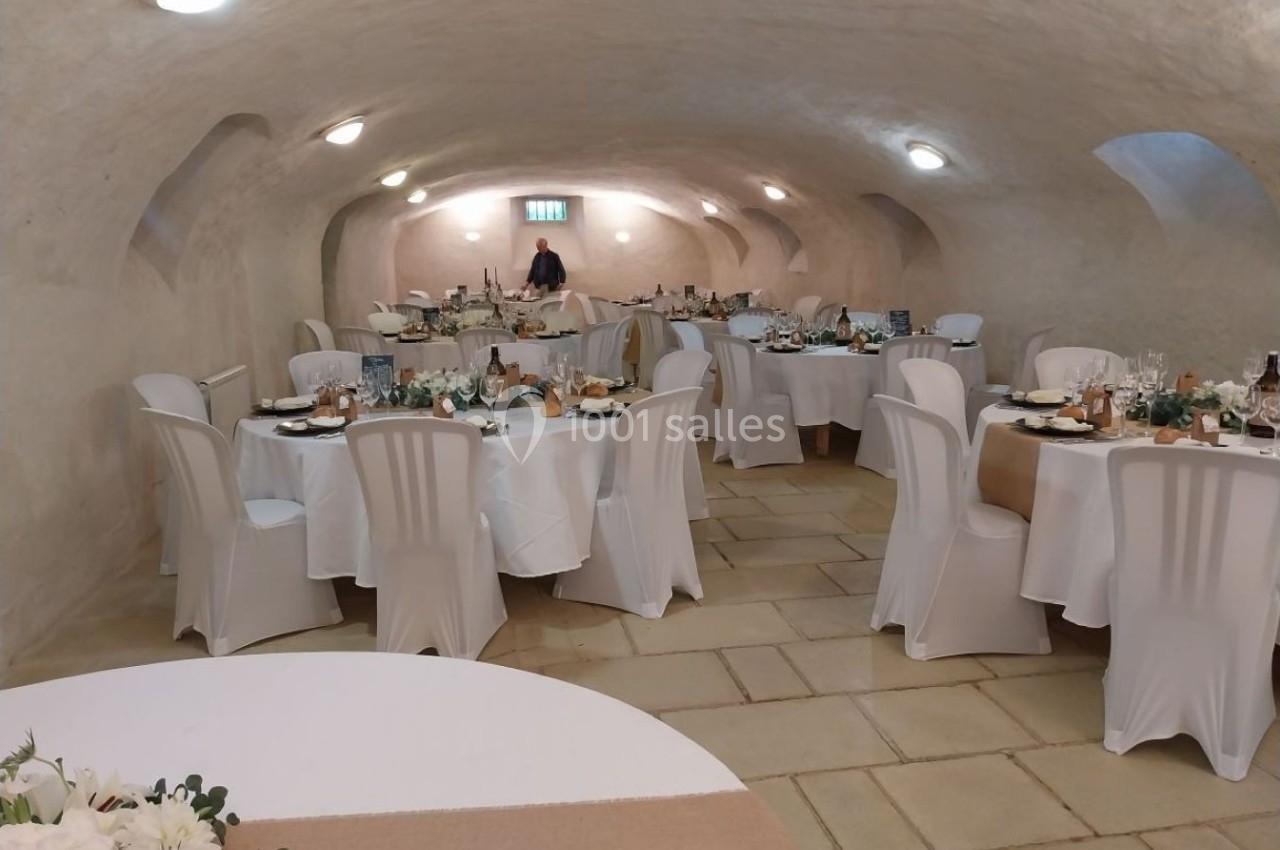 Salle voûtée avec des tables rondes dressées pour un repas, nappes blanches et chaises couvertes.