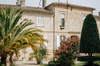 Femme de dos, vêtue d'un chapeau et d'une tenue claire, levant un verre de vin devant une façade en pierre avec volets…