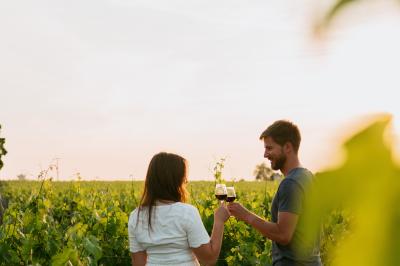 Femme de dos, vêtue d'un chapeau et d'une tenue claire, levant un verre de vin devant une façade en pierre avec volets…