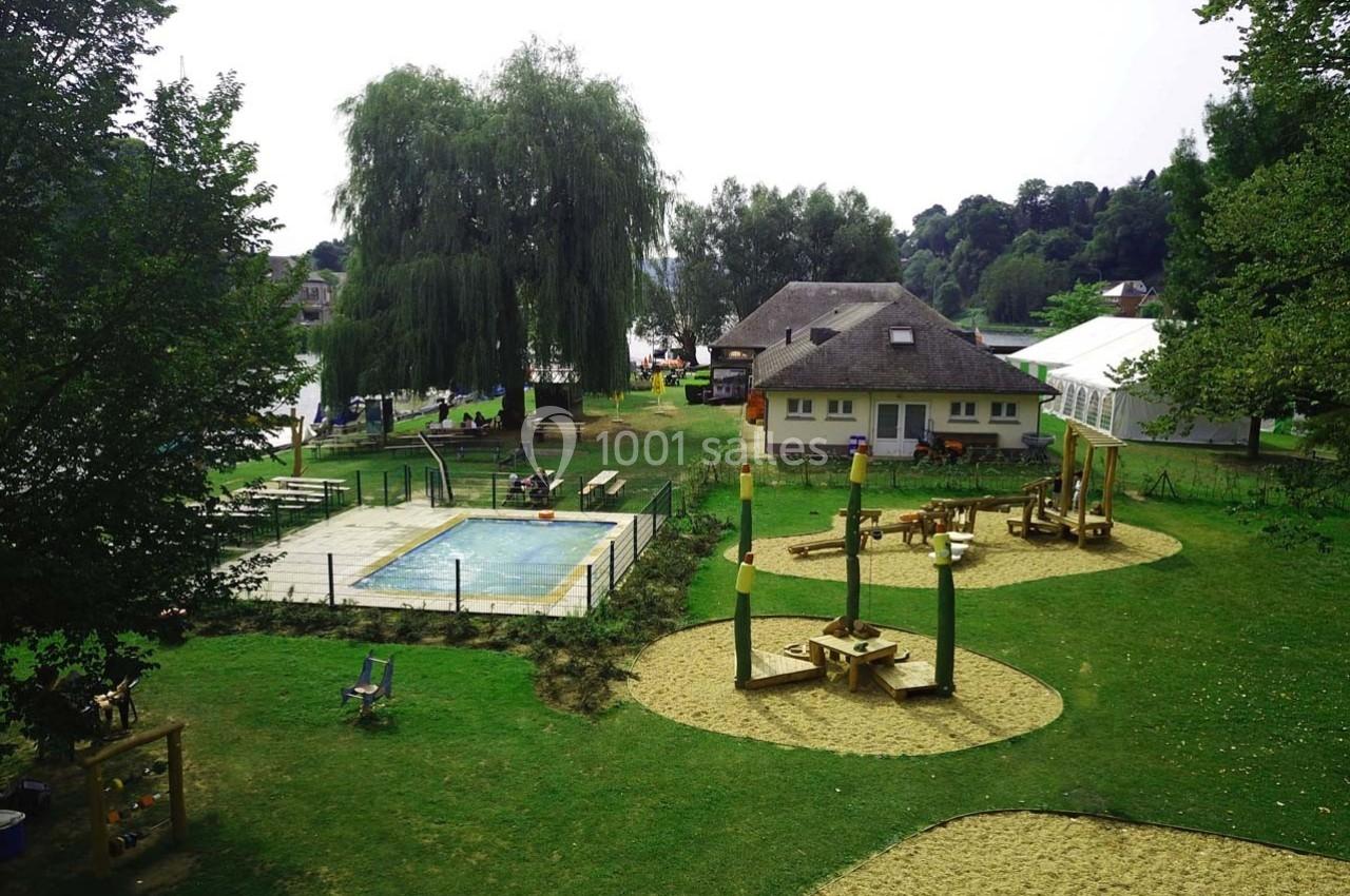 Aire de jeux extérieure avec structures en bois, piscine clôturée et espaces verts entourés d'arbres.