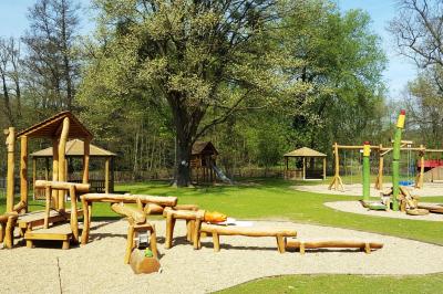 Aire de jeux en bois sur un sol sablé, entourée de verdure et située près d'une rivière et de collines.