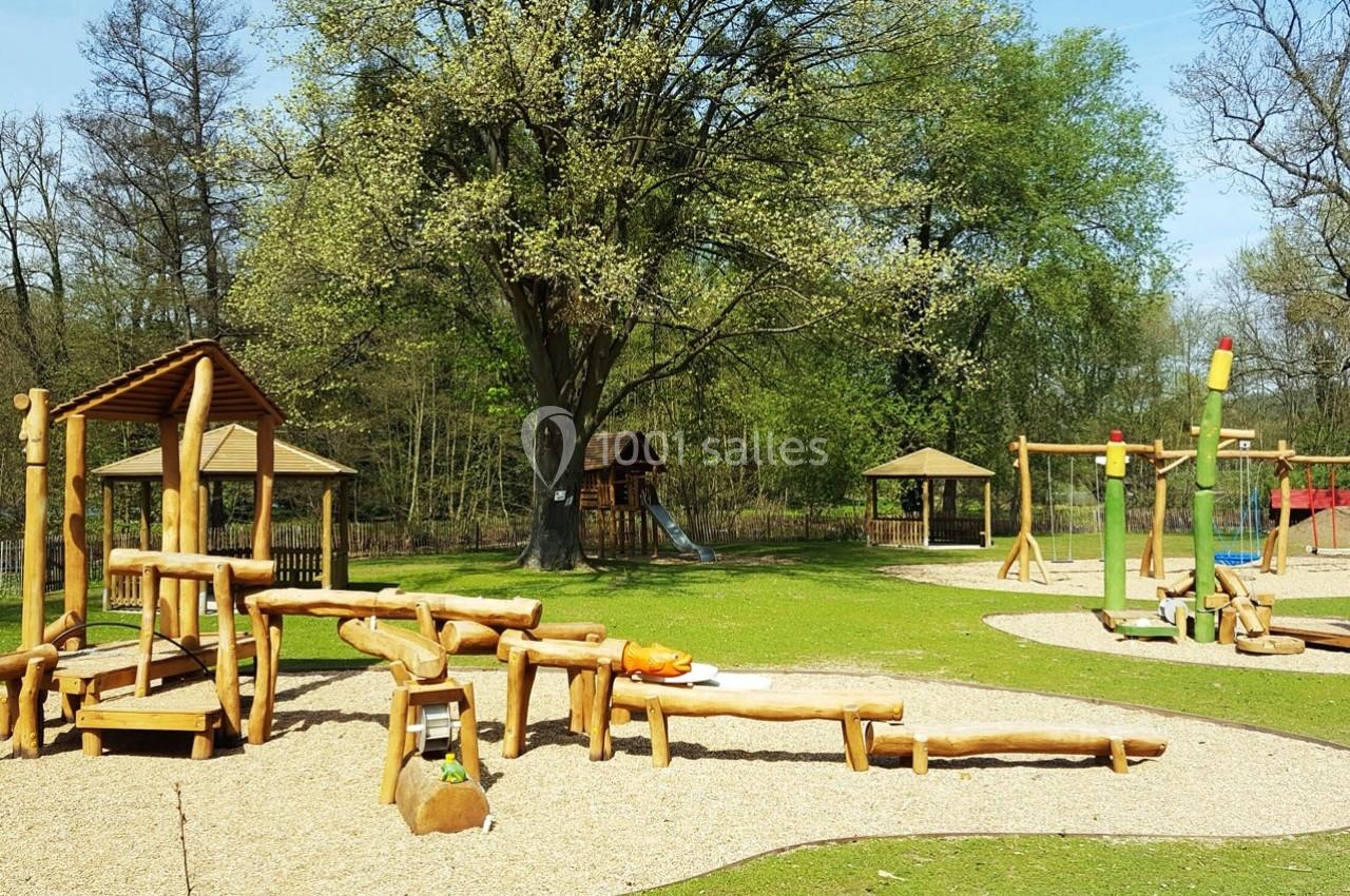 Aire de jeux en bois avec balançoires, toboggan et structures d'escalade, entourée d'herbe et d'arbres.