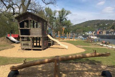 Aire de jeux en bois sur un sol sablé, entourée de verdure et située près d'une rivière et de collines.