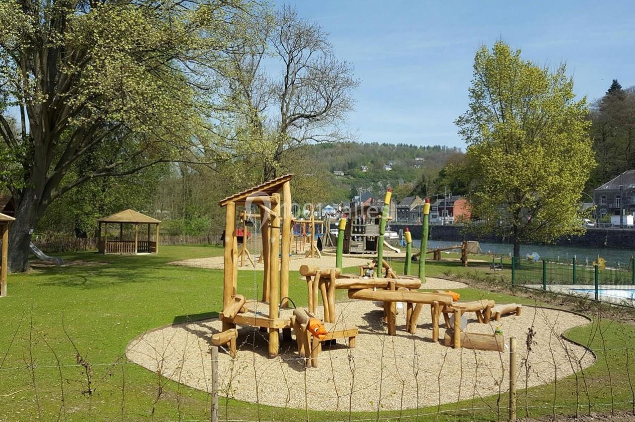 Aire de jeux en bois sur un sol sablé, entourée de verdure et située près d'une rivière et de collines.