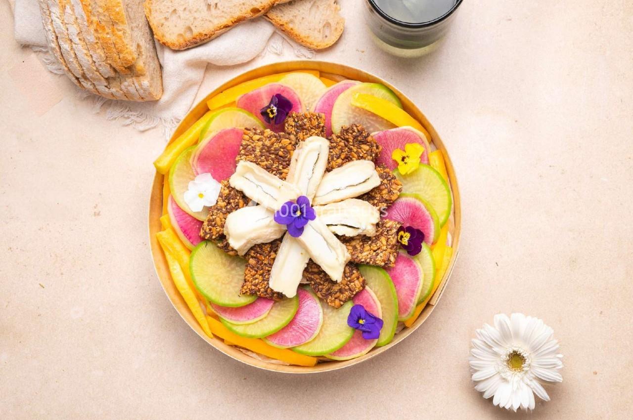 Assiette colorée garnie de légumes tranchés, crackers aux graines, fromage et fleurs comestibles.