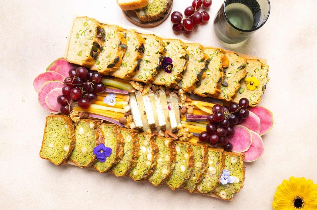 Plateau de pain aux légumes, fromages, fruits frais, noix et fleurs comestibles sur une table claire.