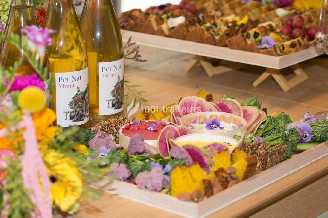 Plateaux de fruits, légumes et fleurs comestibles colorés accompagnés de bouteilles de vin blanc sur une table en bois.