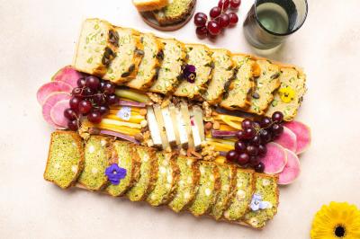 Plateau de pain aux légumes, fromages, fruits frais, noix et fleurs comestibles sur une table claire.