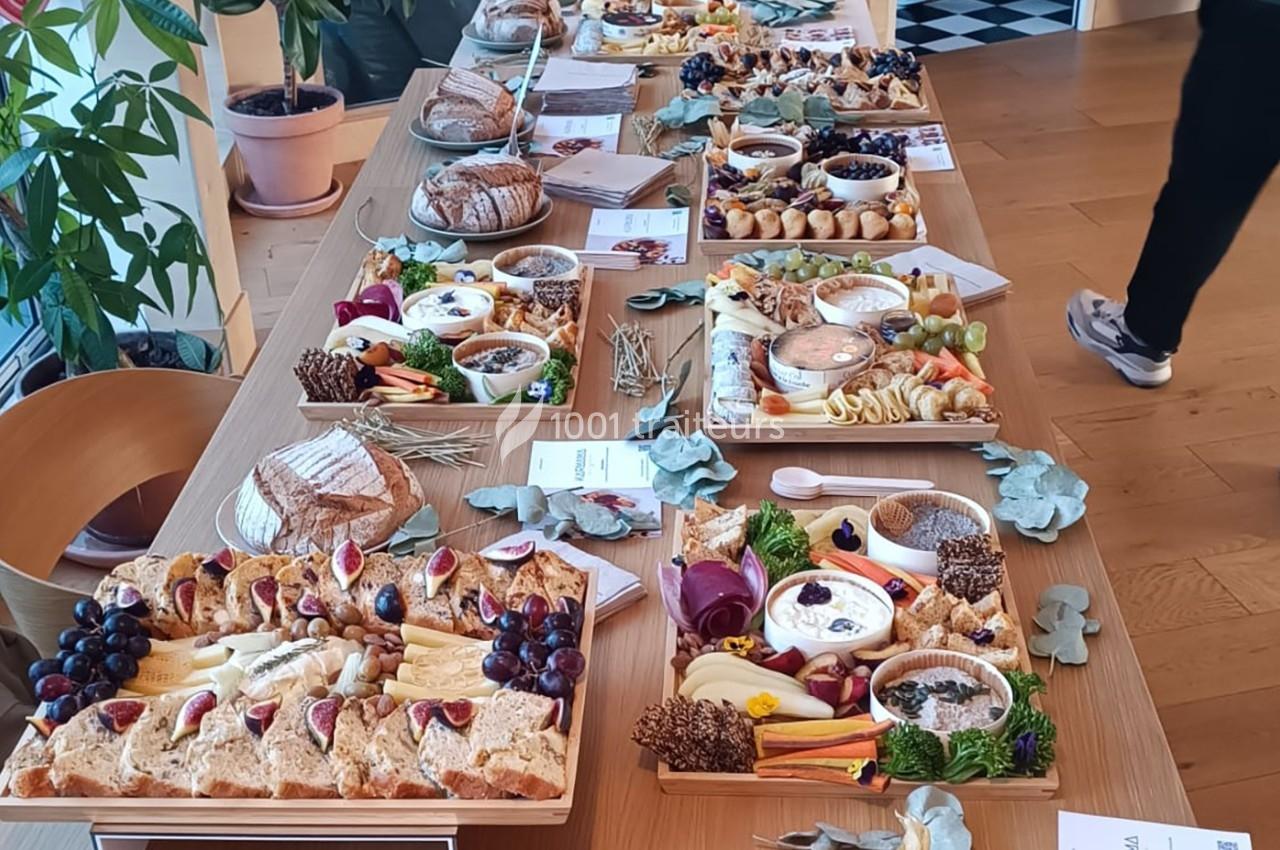 Table garnie de plateaux de charcuterie, fromages, fruits et pains, entourée de plantes et de documents.