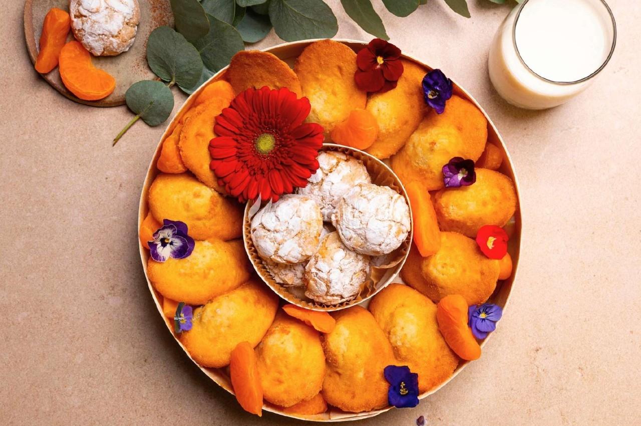 Assortiment de madeleines, biscuits et fleurs comestibles disposés sur un plateau, avec un verre de lait à côté.