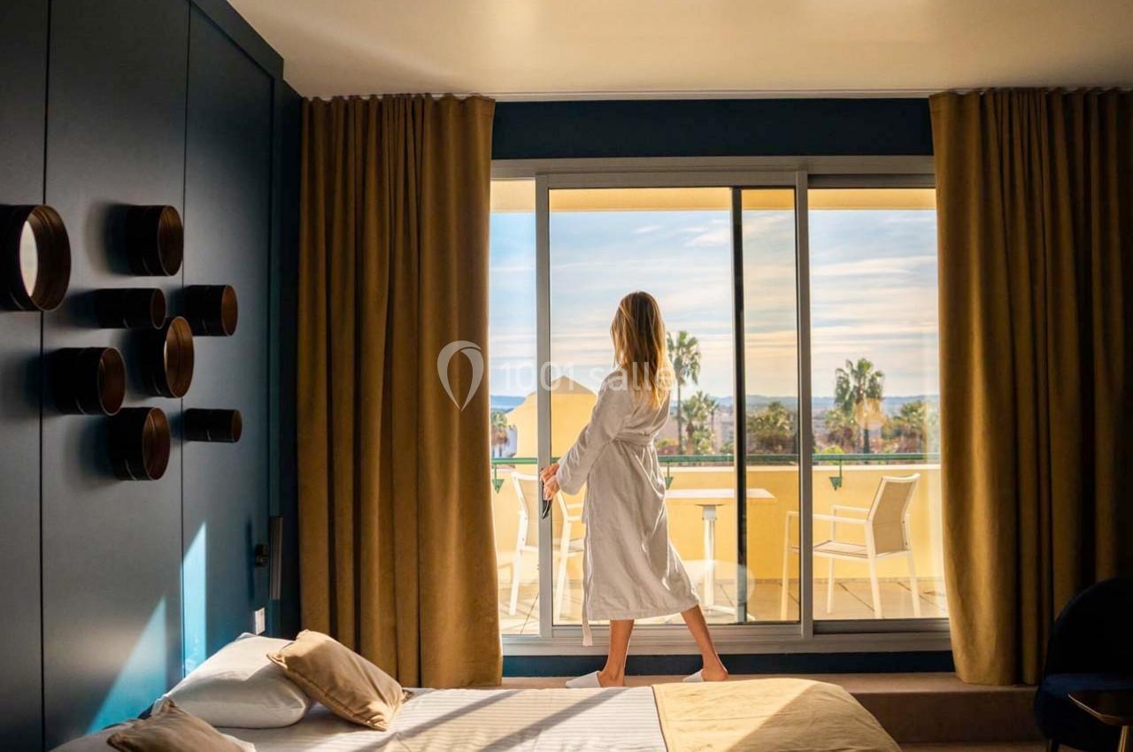 Femme en peignoir regardant à travers une baie vitrée donnant sur une terrasse ensoleillée avec vue sur des palmiers.