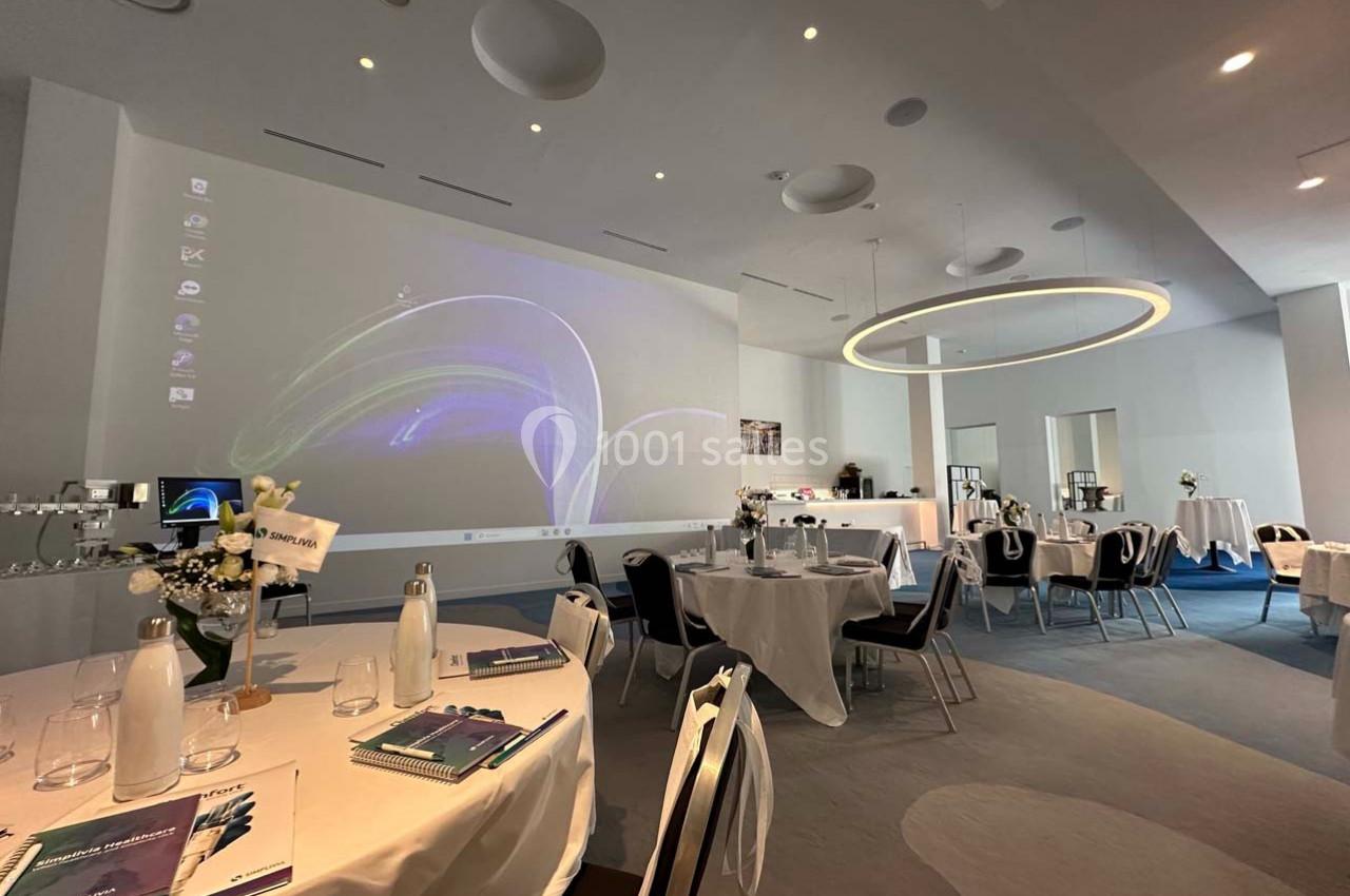 Salle de conférence lumineuse avec tables rondes dressées, projecteur allumé et décor moderne.