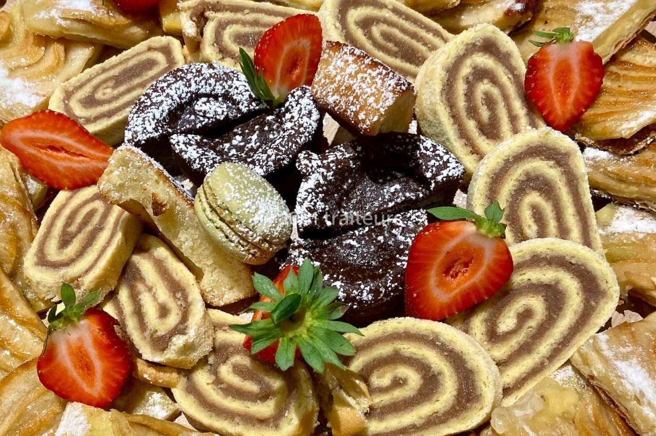 Assortiment de pâtisseries variées avec biscuits roulés, brownies, macarons et fraises fraîches en décoration.
