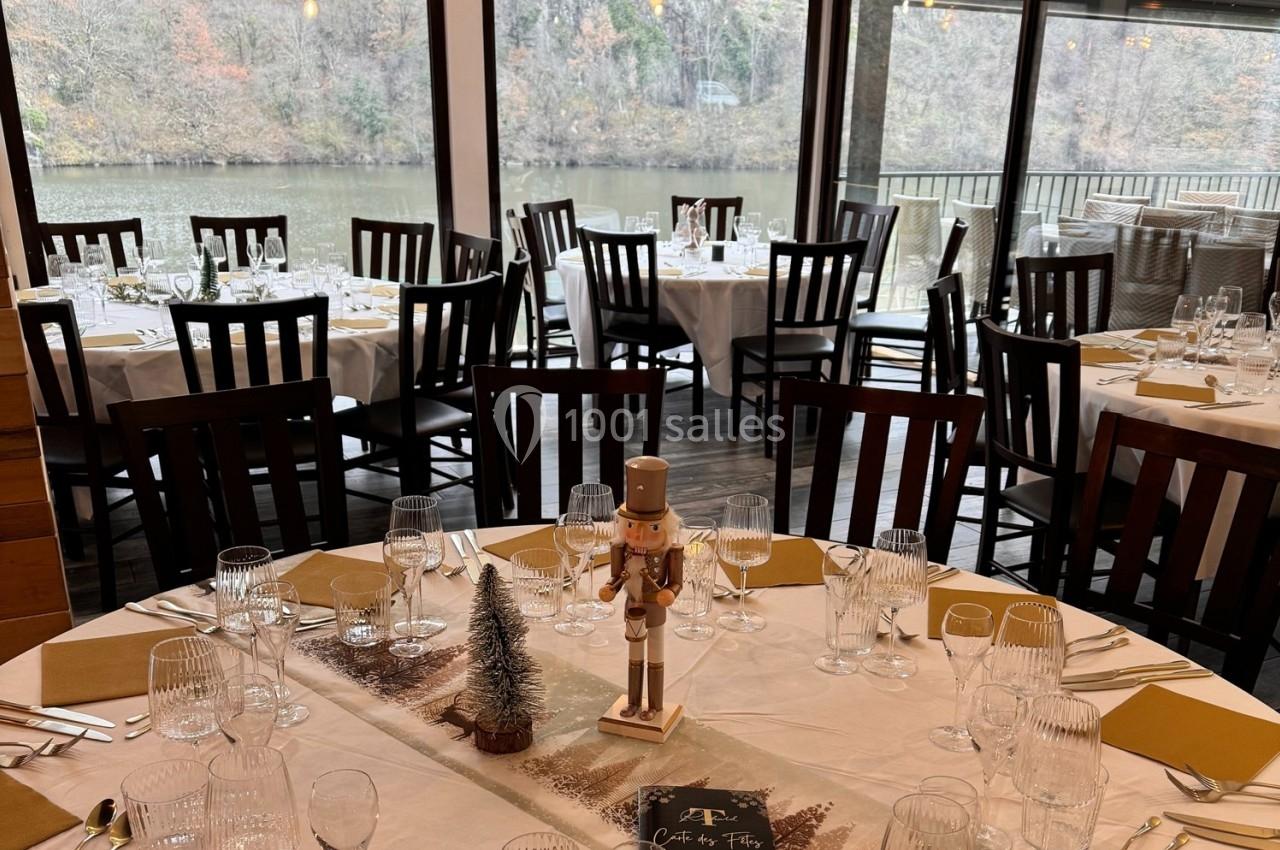 Salle de réception avec tables dressées, nappes blanches, décorations festives et vue sur une rivière à travers de grandes…