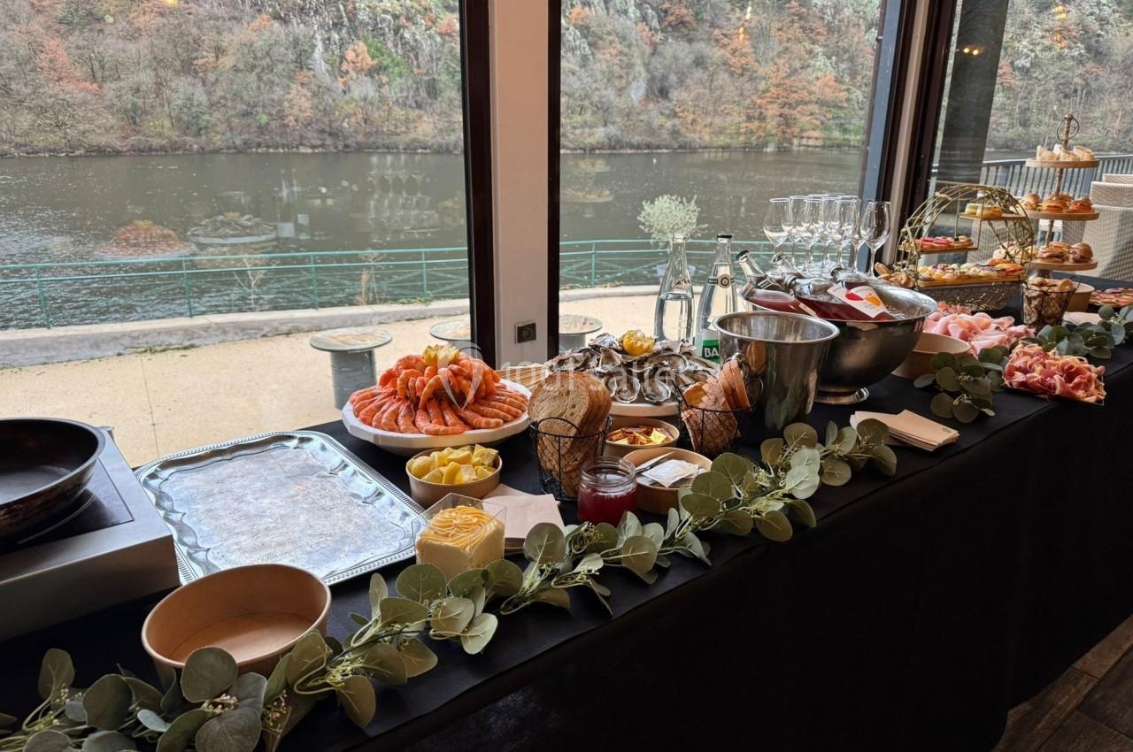 Buffet varié avec fruits de mer, pain, fromages et desserts, disposé près d'une baie vitrée donnant sur une rivière.