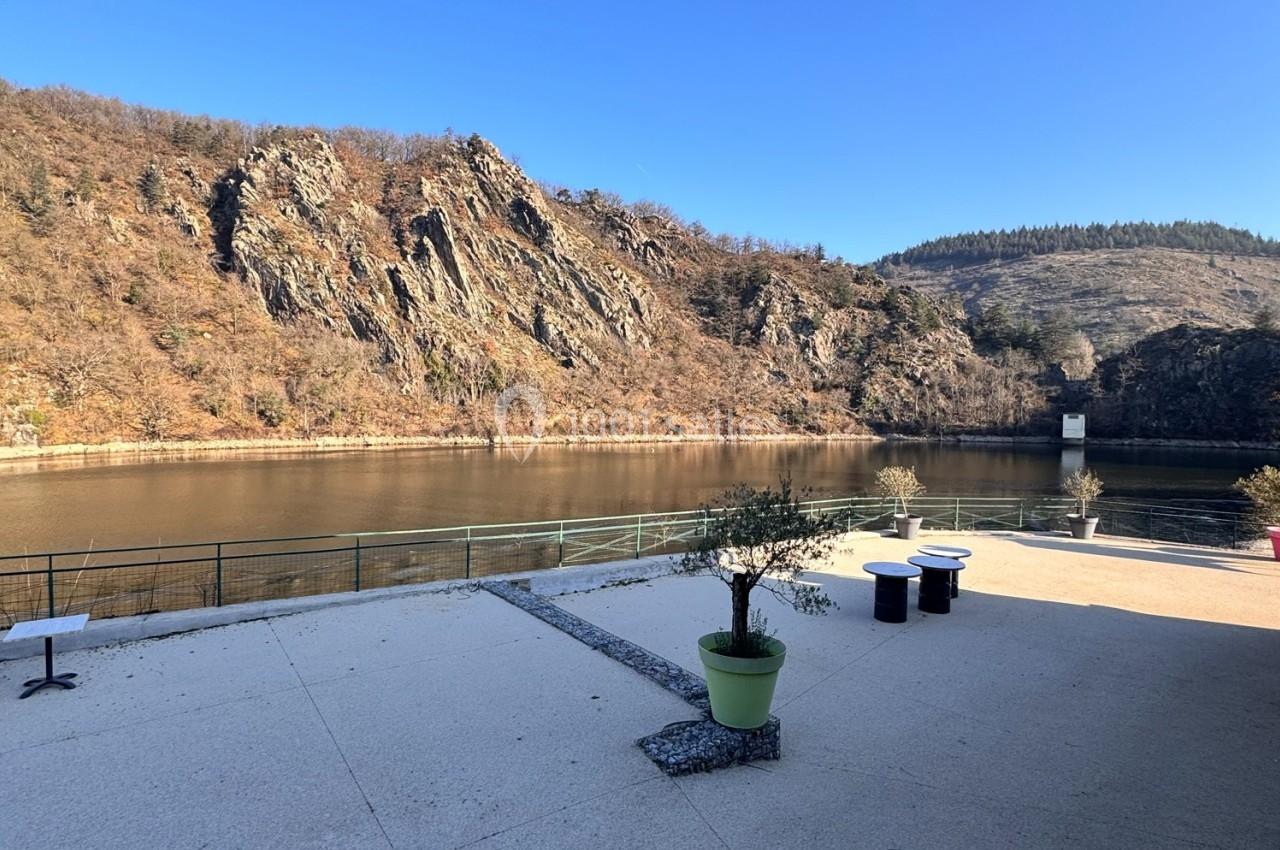 Vue sur un lac entouré de collines rocheuses et boisées, depuis une terrasse avec quelques plantes et mobilier.