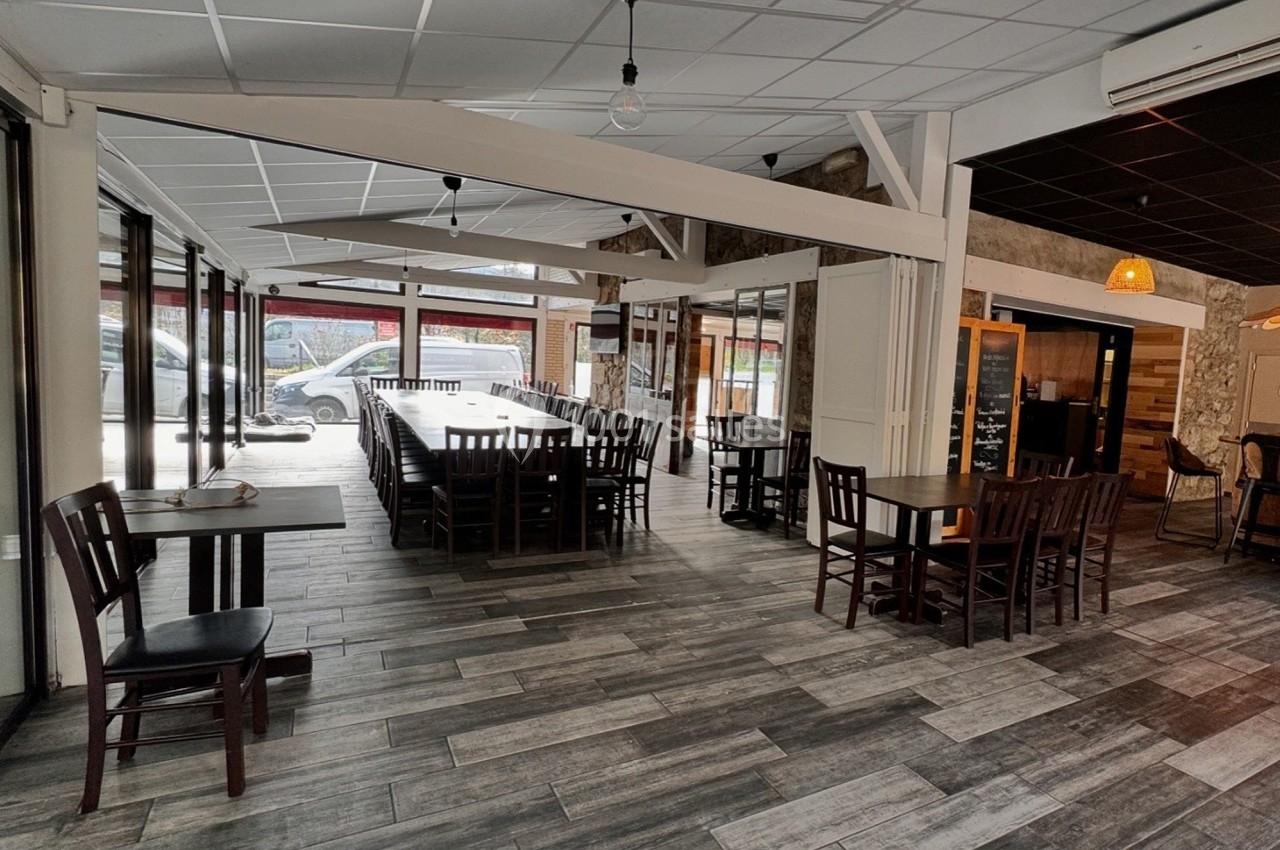 Salle de restaurant lumineuse avec tables en bois sombre, grandes baies vitrées et sol en carrelage gris.