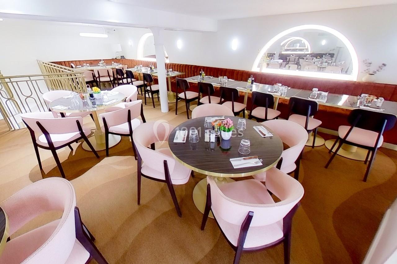 Salle de restaurant lumineuse avec tables rondes en bois sombre, chaises roses et banquettes contre le mur.
