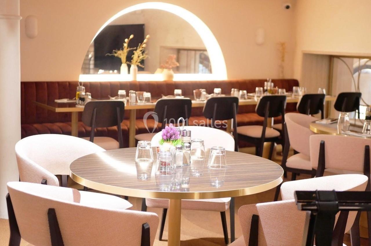Salle de restaurant lumineuse avec des tables rondes, des chaises rembourrées et une banquette en fond sous un miroir…