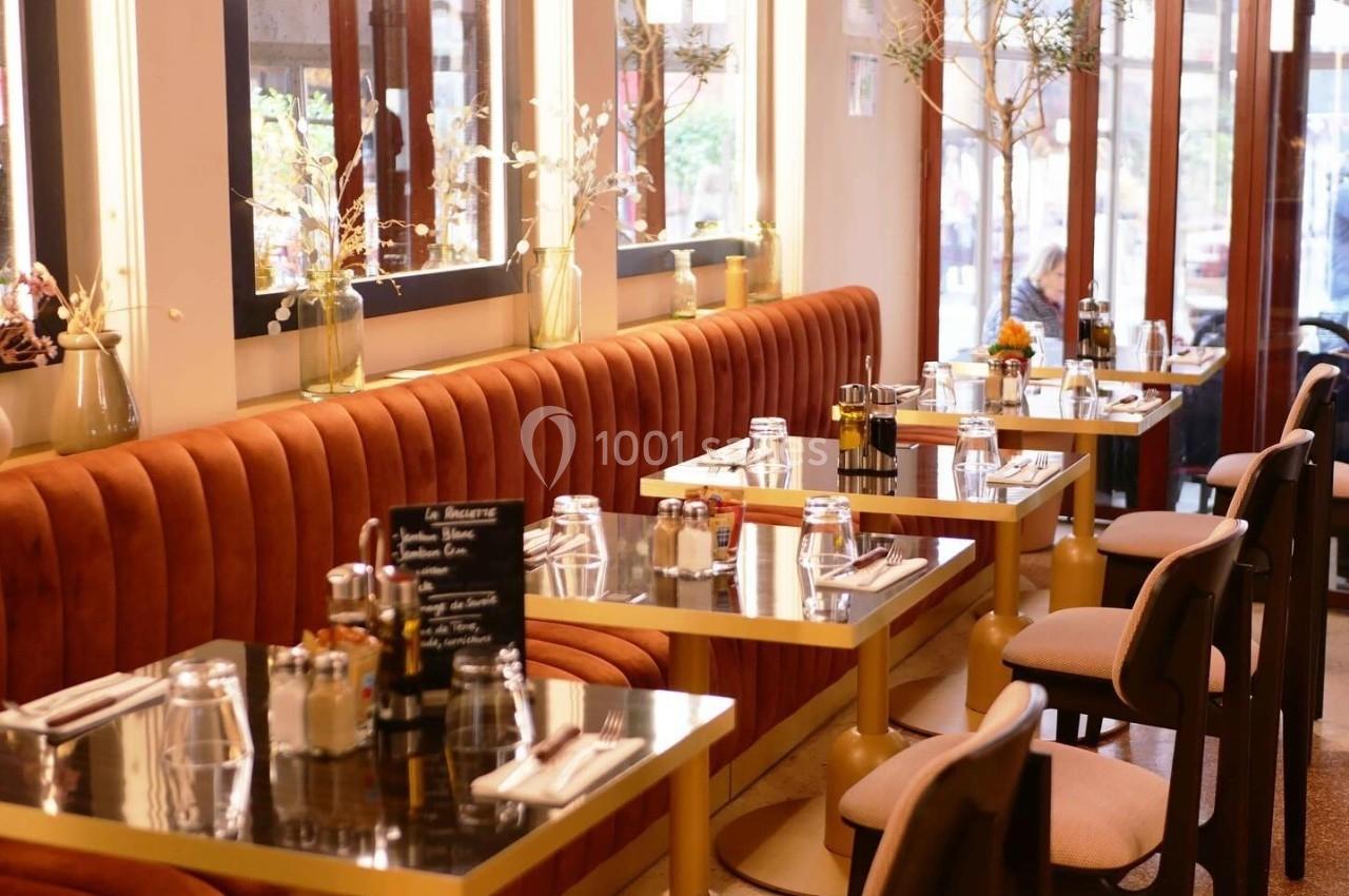 Salle de restaurant avec tables dressées, banquettes en velours orange et grandes baies vitrées laissant entrer la lumière.