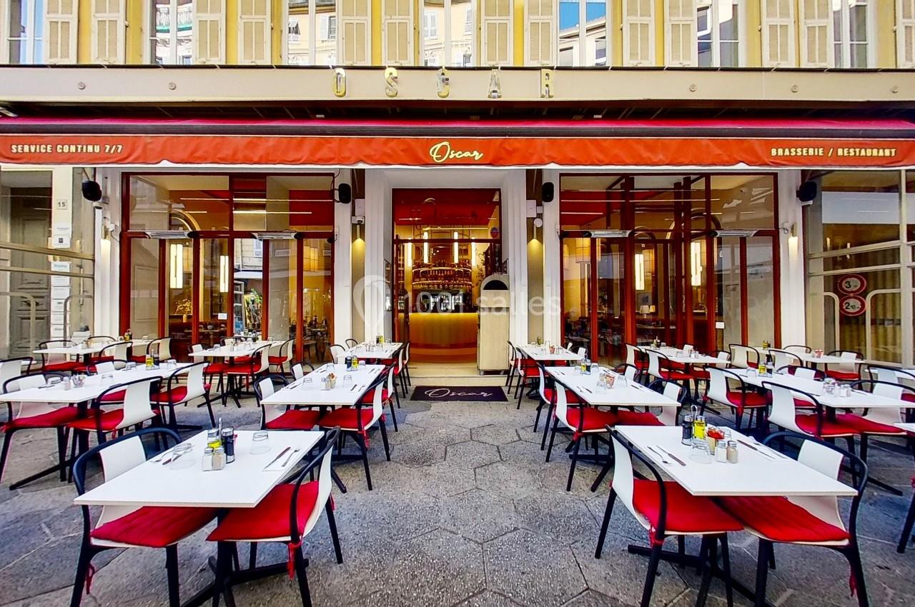 Terrasse d'une brasserie avec des tables et chaises rouges et blanches, située devant une façade vitrée et un auvent rouge.