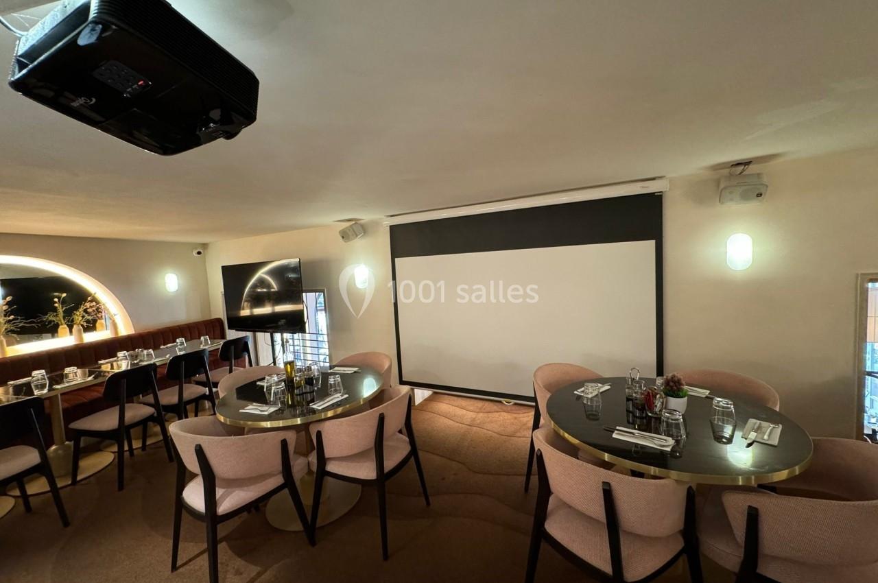 Salle de réunion avec tables rondes, chaises roses, écran de projection et lumière naturelle.