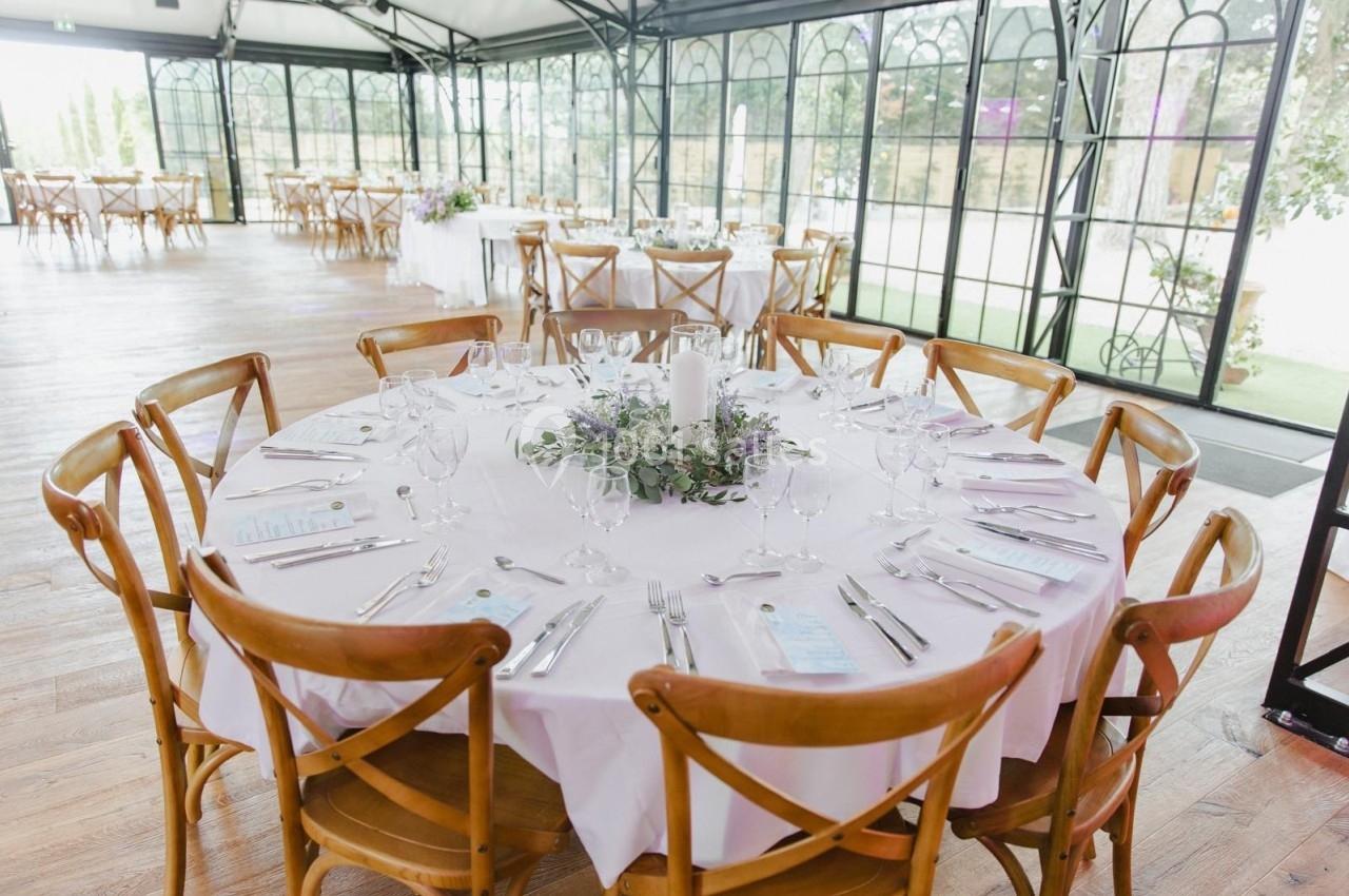 Table ronde dressée avec nappes blanches, couverts, verres et centre de table floral dans une salle lumineuse avec baies…