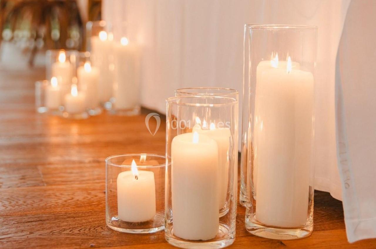 Bougies blanches allumées dans des photophores en verre, disposées sur un sol en bois près d'un rideau clair.