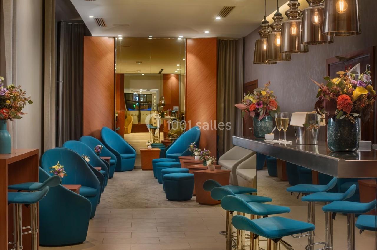 Salon moderne avec fauteuils bleus, tables basses, bar avec tabourets et éclairage suspendu, décoré de fleurs.