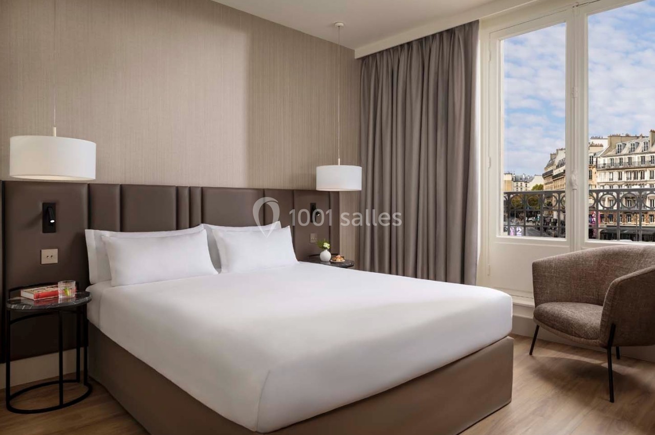 Chambre d'hôtel lumineuse avec un lit double, deux lampes suspendues, un fauteuil et une fenêtre donnant sur des immeubles…