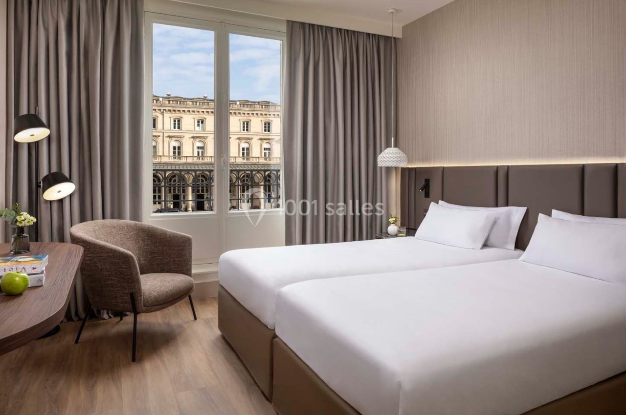 Chambre d'hôtel lumineuse avec deux lits simples, un bureau, un fauteuil et une grande fenêtre donnant sur un bâtiment…