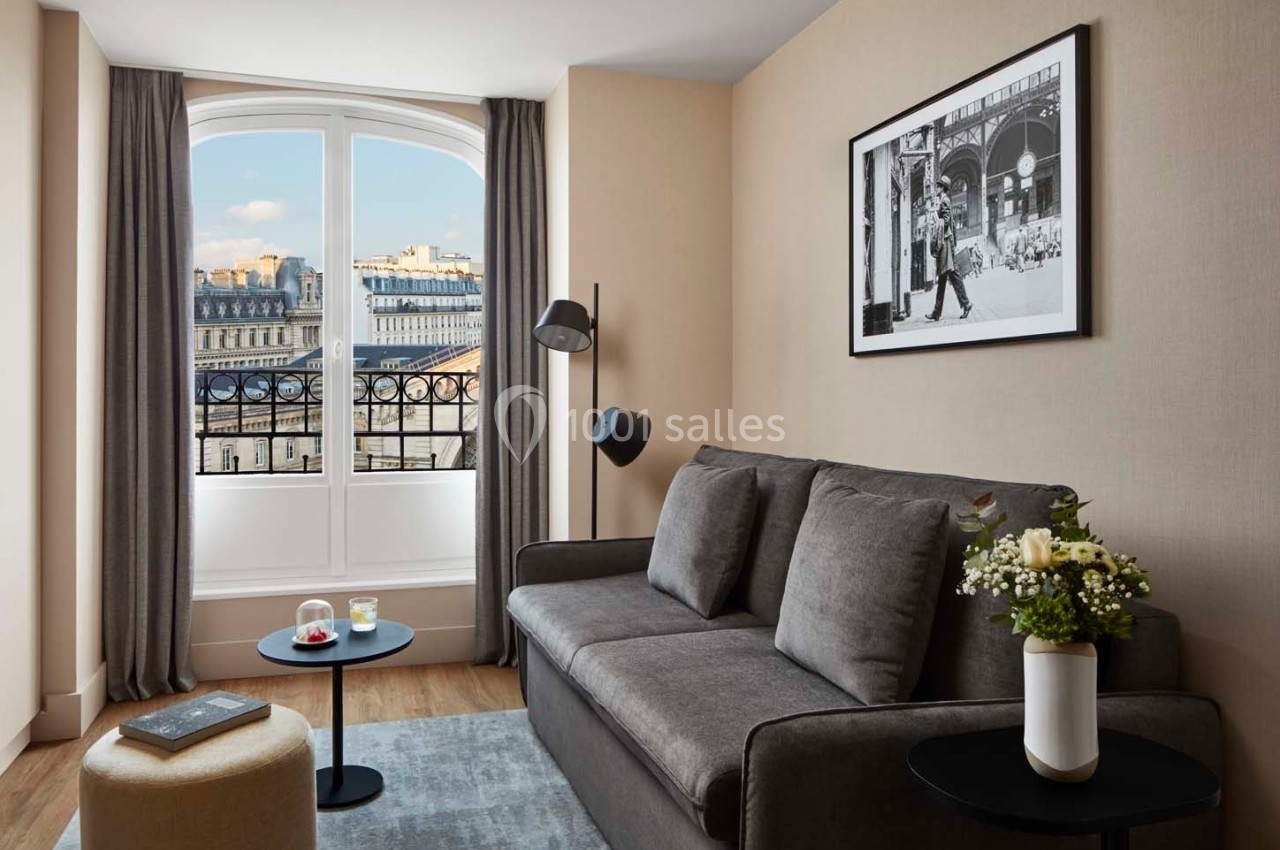 Salon lumineux avec canapé gris, table basse, décoration florale et grande fenêtre offrant une vue sur des bâtiments…