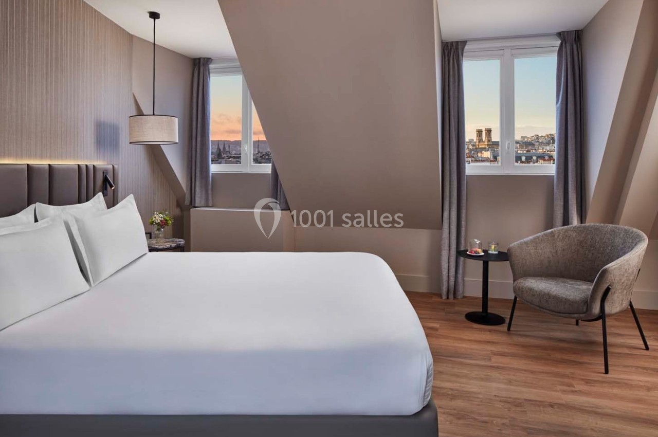 Chambre moderne avec lit double, fauteuil, table basse et vue sur la ville à travers deux fenêtres mansardées.