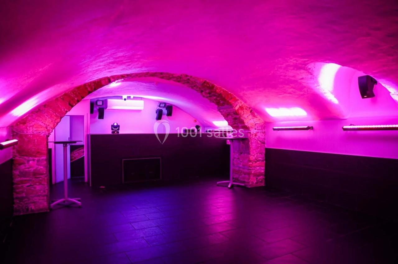 Salle voûtée éclairée par des lumières roses, avec murs en pierre et sol en carrelage sombre.