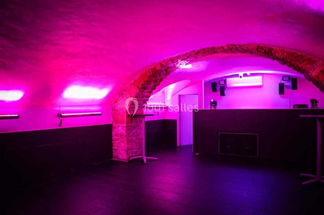 Salle voûtée en pierre avec éclairage rose, équipée de tables hautes et d'enceintes fixées aux murs.