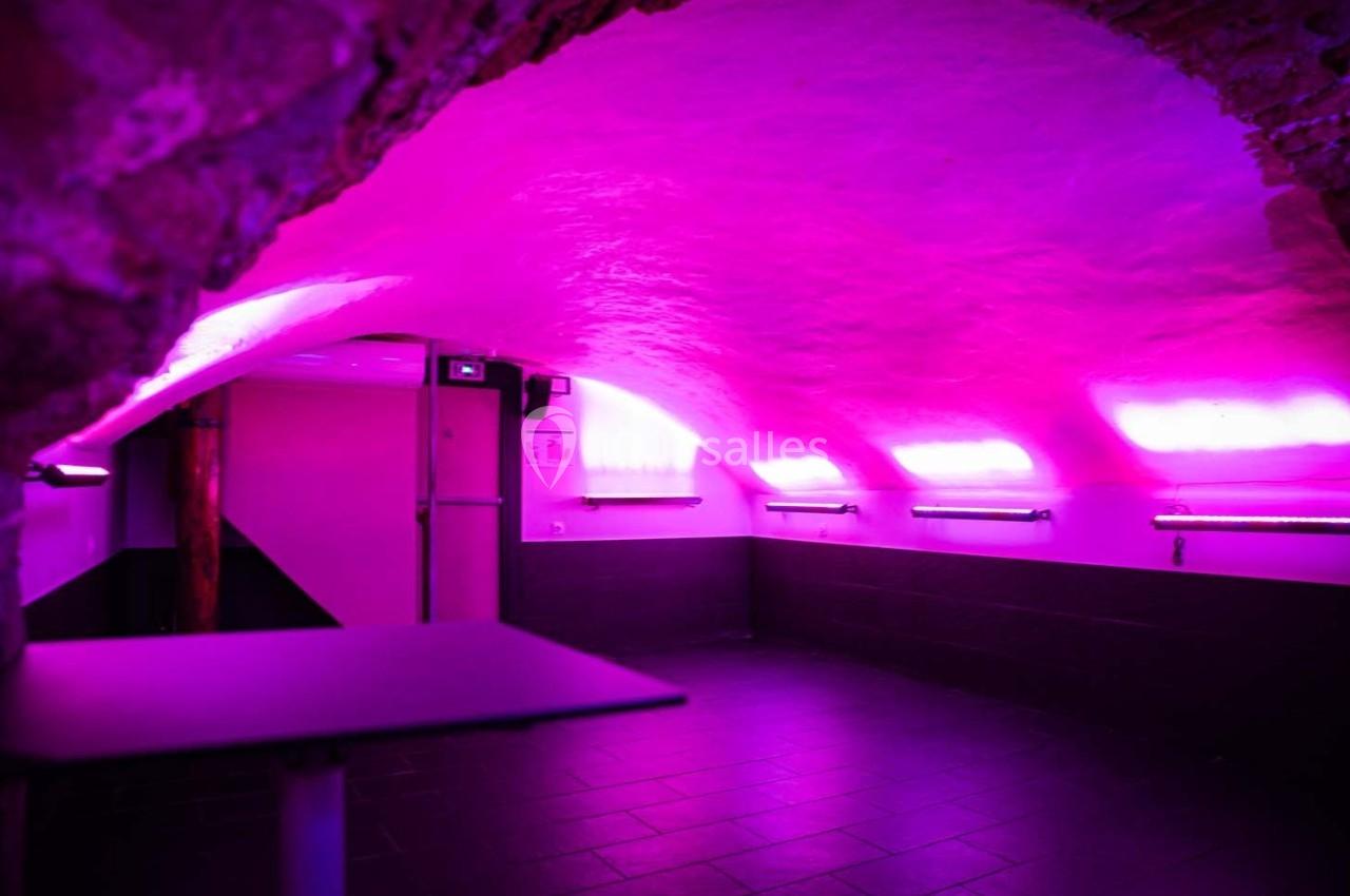 Salle voûtée éclairée par des néons roses, avec murs en pierre et sol en carrelage sombre.