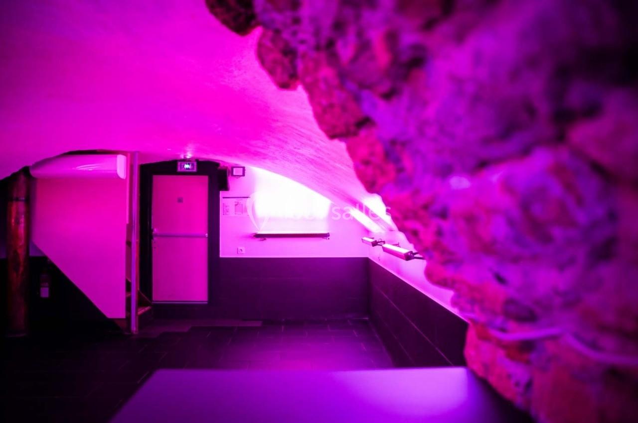 Couloir voûté en pierre éclairé par une lumière rose, avec une porte au fond et des murs blancs.