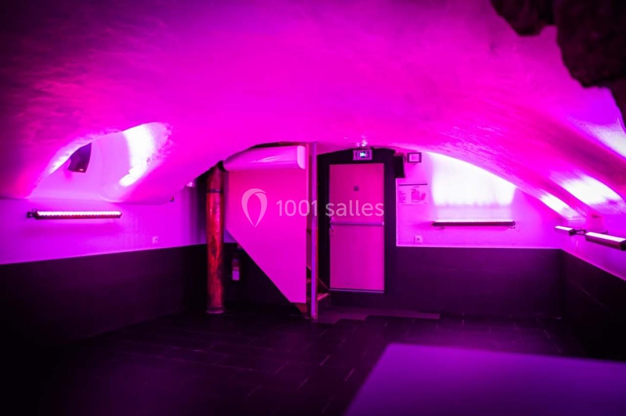 Salle voûtée éclairée par une lumière rose, avec une porte au fond et des murs en pierre peints.