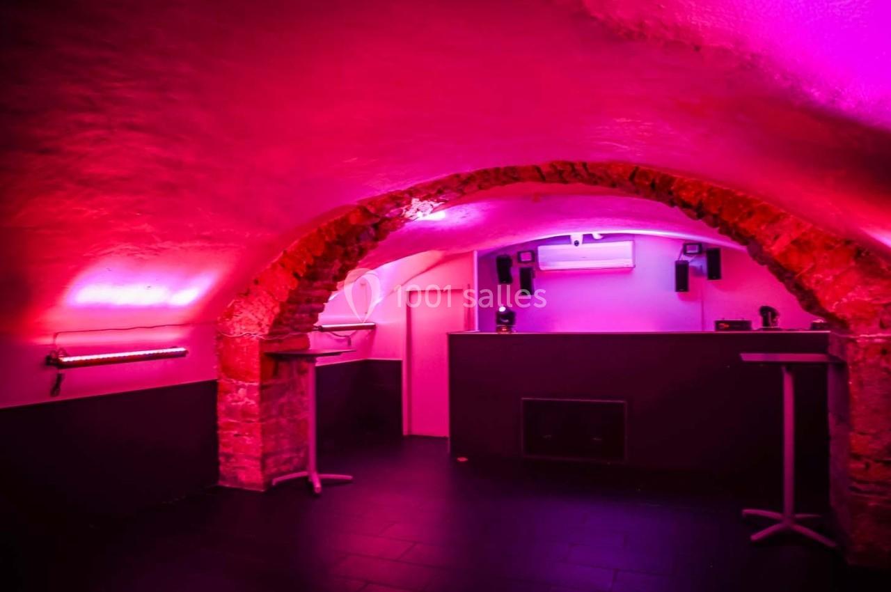 Salle voûtée en pierre avec éclairage rose, tables hautes et un comptoir au fond.