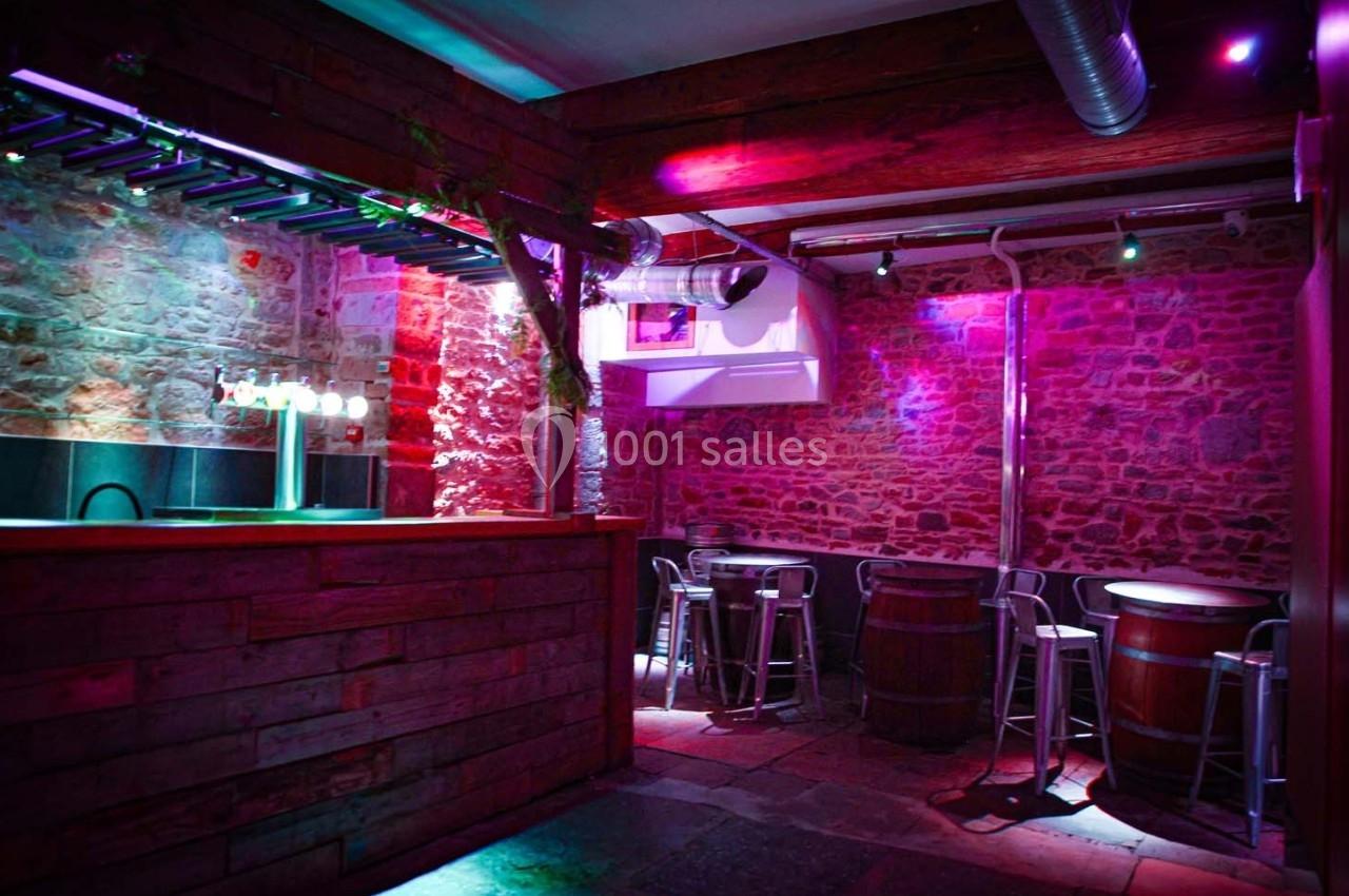Bar en bois avec tabourets et tonneaux dans une pièce en pierre, éclairée par des lumières colorées.