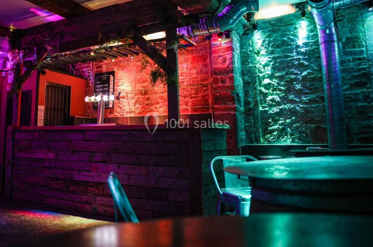 Bar en bois avec éclairage coloré, murs en pierre apparente et tables métalliques dans une ambiance tamisée.