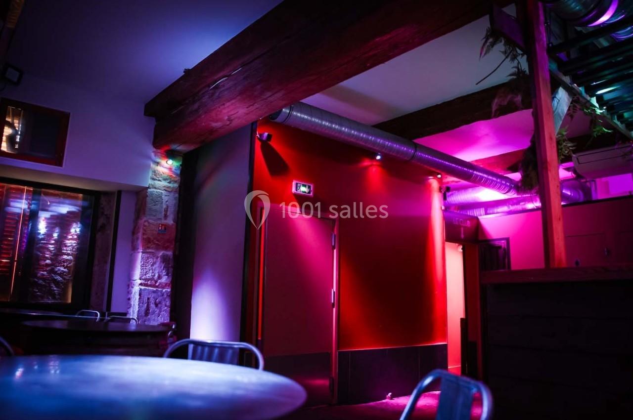 Salle intérieure avec murs rouges, éclairage tamisé aux tons violets et bleus, poutres apparentes et mobilier métallique.