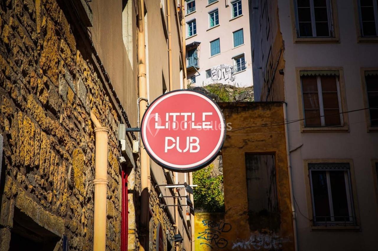 Enseigne ronde rouge avec l'inscription ’Little Pub’, accrochée à une façade dans une rue bordée de bâtiments anciens.