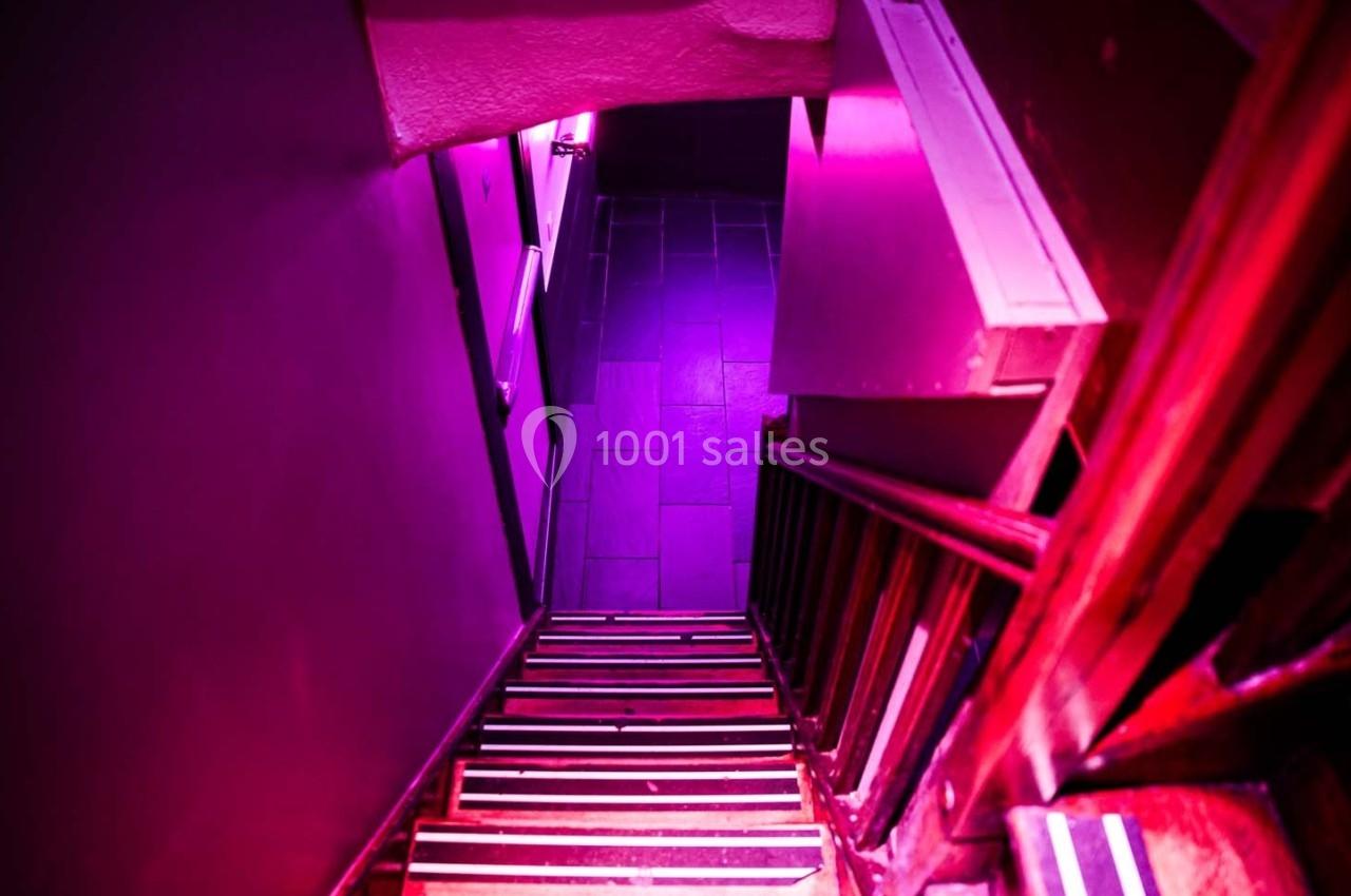 Escalier étroit en bois éclairé par une lumière rose, menant à un couloir sombre avec un sol carrelé.