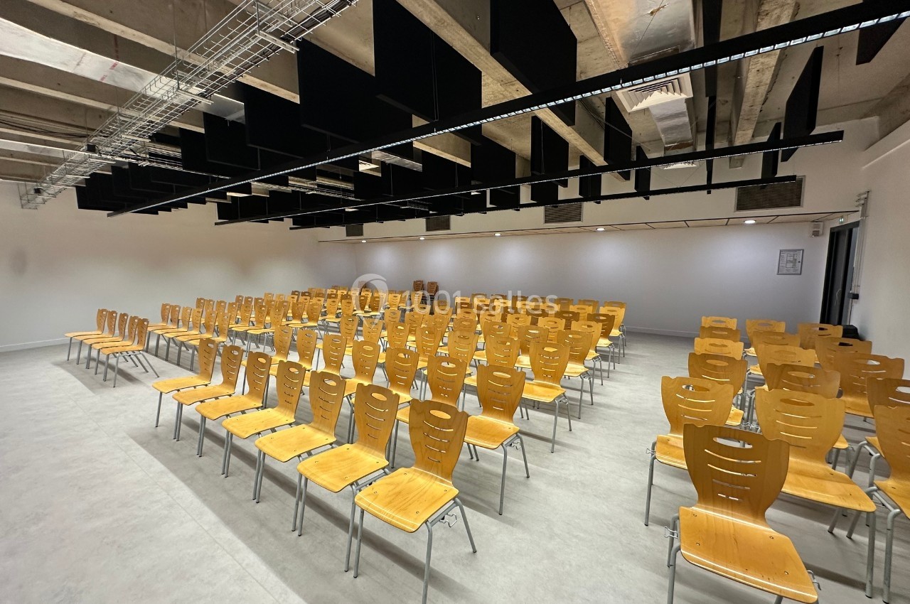 Salle de conférence vide avec des rangées de chaises en bois alignées sur un sol clair, sous un plafond industriel.