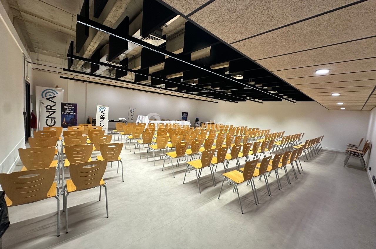 Salle de conférence vide avec rangées de chaises en bois alignées, plafond avec poutres noires et éclairage intégré.