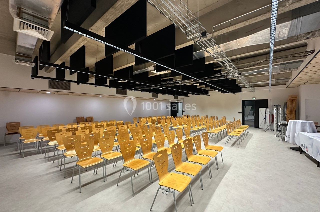 Salle de conférence vide avec des rangées de chaises en bois, plafond industriel et éclairage suspendu.