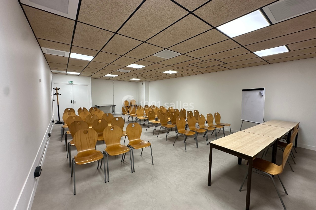 Salle de réunion avec rangées de chaises en bois, tables à l'avant et un tableau blanc sur trépied.