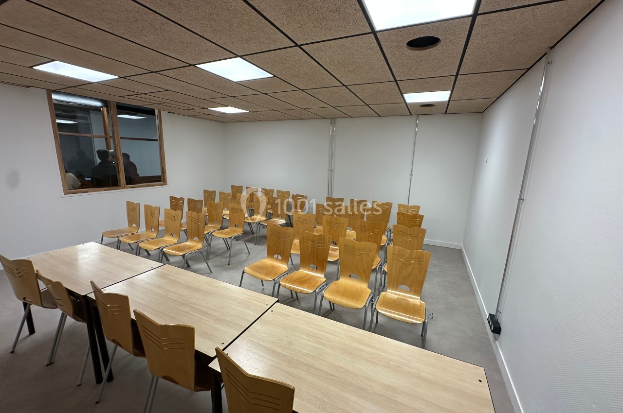 Salle de réunion avec des chaises en bois alignées devant des tables, éclairée par des plafonniers et une fenêtre.