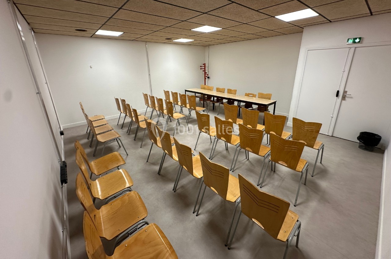Salle de réunion avec des rangées de chaises en bois disposées face à une table et un tableau blanc.